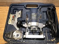 Top-down view of blue and silver Mastercraft plunge router in molded plastic carrying case with attached black power cord, metal plunge base, and metal mounting plate.