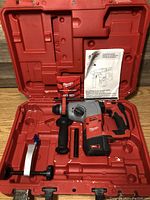Open red plastic case showing drill in molded compartment, side handle, depth rod, manual and accessory packet.