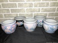 Five ceramic round planters with floral designs arranged on a dark surface against a brick background