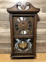 Front view of Sabre pendulum clock showing wood case, Roman numeral dial, and etched lower glass panel with pendulum visible.