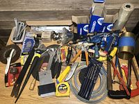 The photo shows a large variety of tools and hardware items arranged on a wooden surface, including several wrenches, a measuring tape, brushes, and packaged clamps.