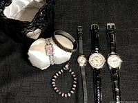 Four women's watches arranged alongside two bracelets on a black fabric background. One watch is Guess brand with pink dial on silver link bracelet, one bracelet is black and gold metal bangle with leopard print, and the other a purple beaded stretch bracelet.