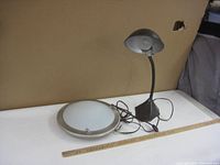 Two lamps shown together: a cast iron desk lamp with gooseneck arm and a round ceiling light fixture with frosted glass and metal trim on a table.