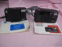 Two vintage cameras with their original manuals laid on a pink background. Left camera is Minolta AF-S and right camera is Pentax Zoom 90 Super.