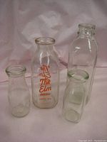 Four antique glass milk bottles displayed on a pink fabric backdrop, one bottle has orange printed text 'The Elm Dairy, Leroy, N.Y.'