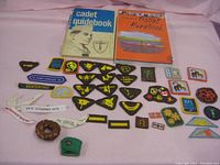 Photo showing Cadet Guidebook, Scout Handbook, and a variety of scouting patches and badges arranged on a pink surface.