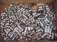 Close-up photo showing a large assortment of metal sockets in a box, mainly cylindrical shapes with hexagonal internal shapes for gripping nuts and bolts.