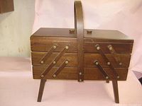 Closed view of vintage wood sewing basket showing dark brown finish, hinged lid, accordion style sides with brass hardware, four legs, and single arched handle.
