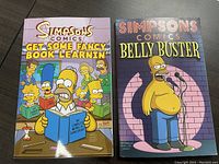 Front cover of the two comic paperbacks, one titled 'Get Some Fancy Book Learnin'' featuring Simpsons characters in a library, the other titled 'Belly Buster' showing Homer on stage.