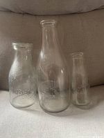 Three clear glass vintage milk bottles with embossed names displayed on a beige fabric surface.