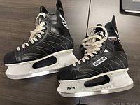 Pair of black Bauer TUIK Impact 100 Pro hockey skates with white laces and blade holders shown from the side.