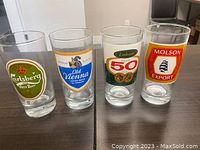 Set of four vintage beer glasses displayed on dark table showing front logos: Carlsberg, Old Vienna, Labatt, Molson Export.