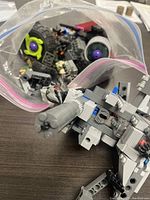 Bag of assorted vintage LEGO parts featuring wheels, bricks, and mechanical elements in grey, black, and purple.