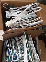 Two cardboard boxes filled with assorted white plastic and metal clothing hangers.