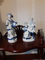 Photo of two porcelain figurines on a dark wooden surface, showing the single female figurine with a hat and the pair figurine with a man and woman dressed in blue and white.