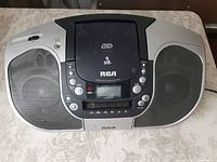 Front view of RCA portable boombox detailing speakers, control buttons, CD player cover, cassette deck, and display screen.