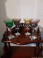 Four coloured wine glasses displayed on a wooden table with carved details. From left to right: green, amber, yellow, and purple glasses. Each has textured glass detail and silver-tone stems and bases.