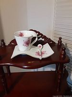 White porcelain tea cup with pink floral design displayed on matching square saucer with matching spoon on wooden table.