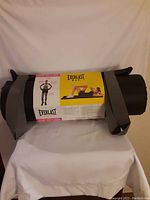Rolled black nylon-covered foam exercise mat with grey carrying straps, in packaging showing the Everlast F.I.T. branding and imagery of fitness exercises.