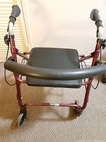 Front view of a burgundy four wheel walker with black padded seat and handles, showing caster wheels and brake handles.