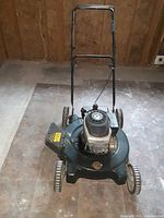 Full view of the black gas lawnmower showing engine, wheels, and handlebar on stone/tile floor inside a room.