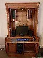 Front view of solid wood armoire desk with both pieces assembled, desktop surface with computer monitor, keyboard, speakers, and telephone in photos. Shows carved details and shelving.