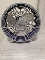 Front view of Sunbeam 9-inch table fan showing three metal blades behind protective grill and Sunbeam logo in center.