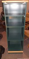 Photo showing front view of a green wooden bookcase with four shelves. The bookcase has a light wood top and bottom trim. Background includes indoor setting with furniture.