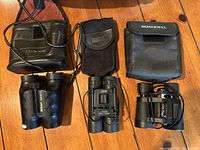 Three pairs of lightweight field binoculars laid on wooden floor with their respective carrying cases: Nikon Travelite III, DRW 10x25, and Bushnell