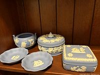 Five Wedgwood blue jasperware pieces displayed on a wooden shelf. Includes two lidded boxes, one handled pot, two small round dishes with cream classical relief decorations. One piece shows damage.