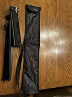 Photo of folded black metal adjustable music stand next to black nylon carry case with shoulder strap lying on wood surface.
