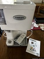 Cuisinart soft serve ice cream maker unit placed on wooden table with instruction manual beside it and some detachable parts assembled in the tray.