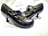 Side angle view showing left and right black leather shoes with distinctive curved heels and Mary Jane straps with silver buckles.