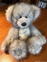 Front view of the handmade mink fur teddy bear sitting upright on a wooden table, showing full body, face, and necklace with name beads.