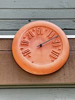Photo of round terracotta outdoor thermometer mounted on wooden wall showing temperature reading at about 85 degrees Fahrenheit.