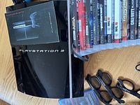 Glossy black PlayStation 3 console with manuals and a corner view showing some accessories and games nearby.