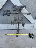 Front view of the entire cast iron weathervane showing cow figure, arrow with fletching and arrowhead, directional arrows, and base with mounting holes, placed near white wooden siding. Tape measure next to it shows width approx 11 inches.