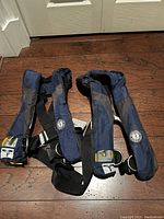 Two navy blue Mustang Survival self-inflating life jackets displayed on floor, showing harness straps and inflation mechanism packs.