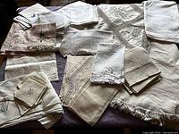 Wide view of assortment of vintage linens including folded tablecloths, embroidered and crocheted pieces, and napkins arranged on a surface.