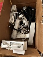 Box containing white Nintendo Wii console, two white remotes, two nunchuks, charging dock, black microphones, and several Wii game cases.