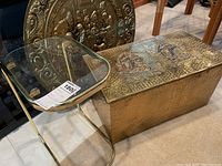 Side view of glass side table with brass frame next to embossed metal chest and round metal wall art