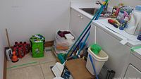 Photo showing broom handles, mop, green and beige cleaning bins, white step stool, various cleaning bottles, and cleaning supplies in a corner with sink.
