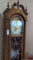Front view of tall wooden grandfather clock showing arched top with finials and ornately decorated clock face with Arabic numerals behind glass door.