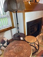 Wood floor lamp with wicker shade, globe-style inner lamp shade, and two side tables: a woven rattan table and a carved wooden Indian side table.
