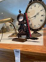 Photo showing two ornate reproduction table clocks with roman numeral dials, a brass desk lamp, and a small metal dustpan on a wooden surface.
