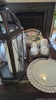 Photo showing black metal lantern with white candle, three glass candle holders with white candles, clock, and decorative mirror on table.