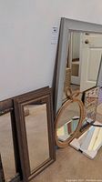 Photo showing six mirrors leaning against a wall, various shapes including large rectangular and oval, different frames like ornate and rope style.