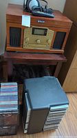Full view of radio CD player atop a small wooden table, with CDs and black plastic CD storage box in front, and Sony headset and remote on top.