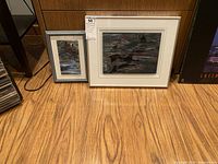 Wide shot showing both framed sandblasted glass artworks side by side on a wood floor.