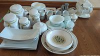 Wide view of the ceramic serveware and storage containers arranged on a wooden floor showing multiple items including jars, plates, bowls, salt and pepper shakers, and a covered terrine.